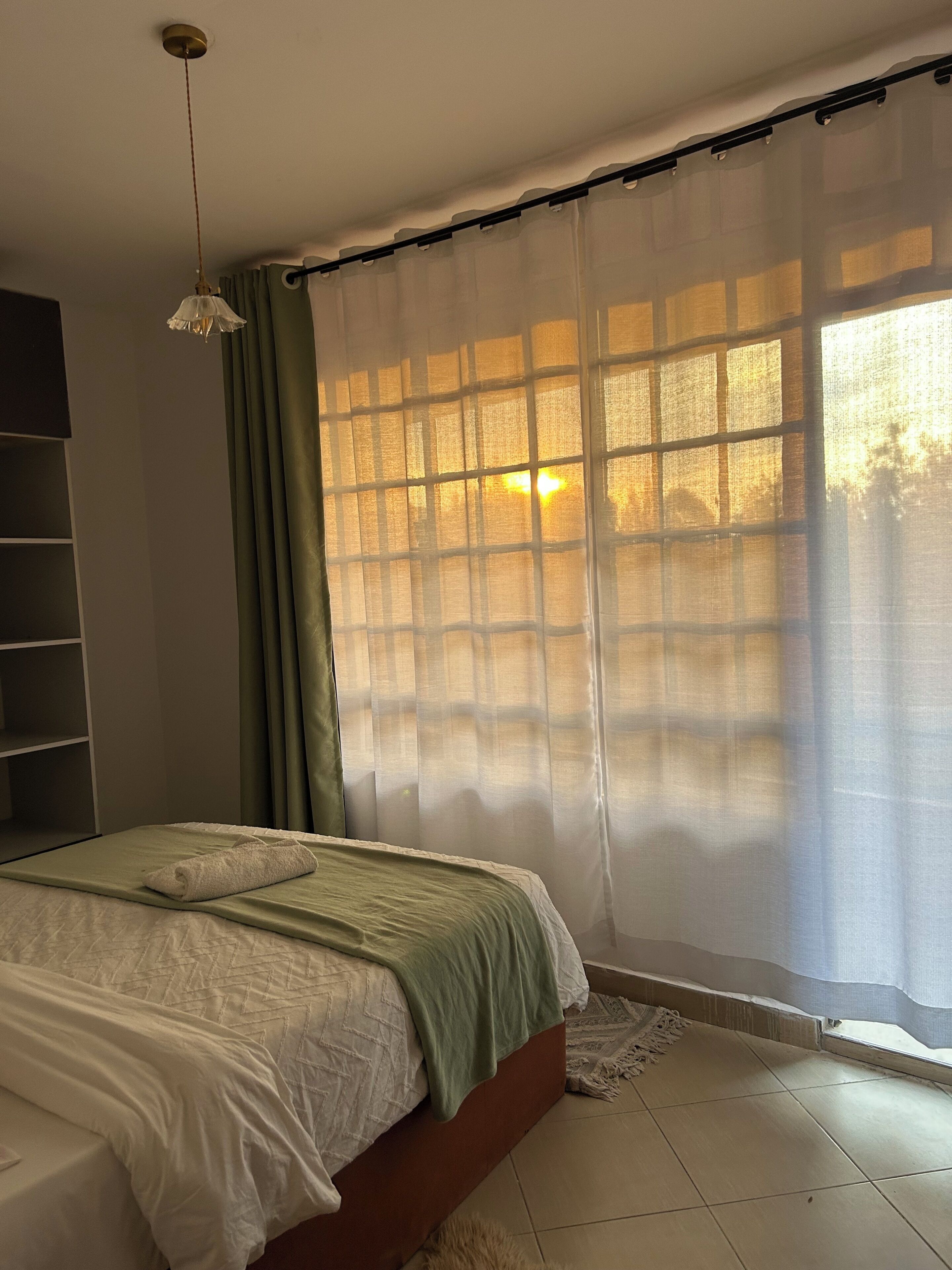 Ein Schlafzimmer mit Bett, einer Hängeleuchte, einem Fenster mit durchsichtigen Vorhängen und Blick ins Freie.