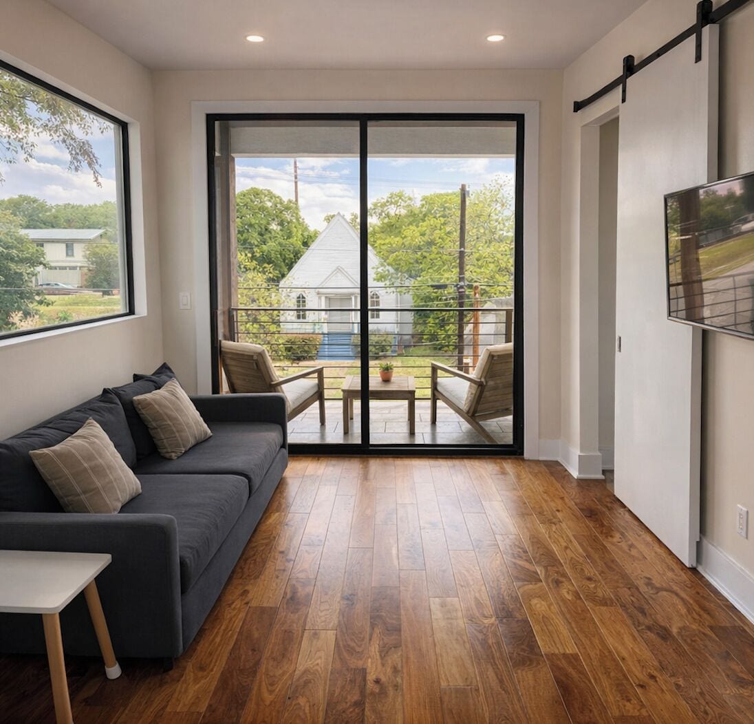 A modern living room with a dark sofa, wooden flooring, and a large sliding glass door leading to an outdoor patio.