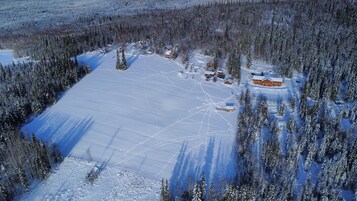 A snow-covered landscape with a large open area, several buildings, and dense forest.