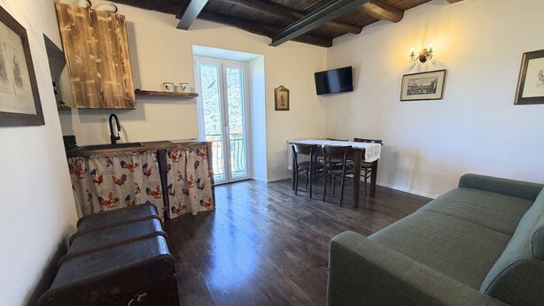 A living room with a green sofa, a wooden table with chairs, a kitchen area with a window, and a wall-mounted TV. - Enchantment of the Lake app Menaggio two-room apartment with bathroom and balcony (Molina)