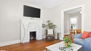 Una sala de estar con chimenea de ladrillo blanco, un televisor de pantalla plana, un sofá azul y un comedor al fondo.