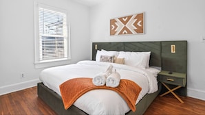 Un dormitorio con cama, mesita de noche, una obra de arte en la pared y una ventana con persianas.