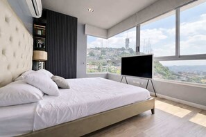A bedroom with a large bed, a flat-screen TV, a window with a city view, and a built-in air conditioning unit. - Stylish Loft, Balcony and City Views with Parking (Guayaquil)