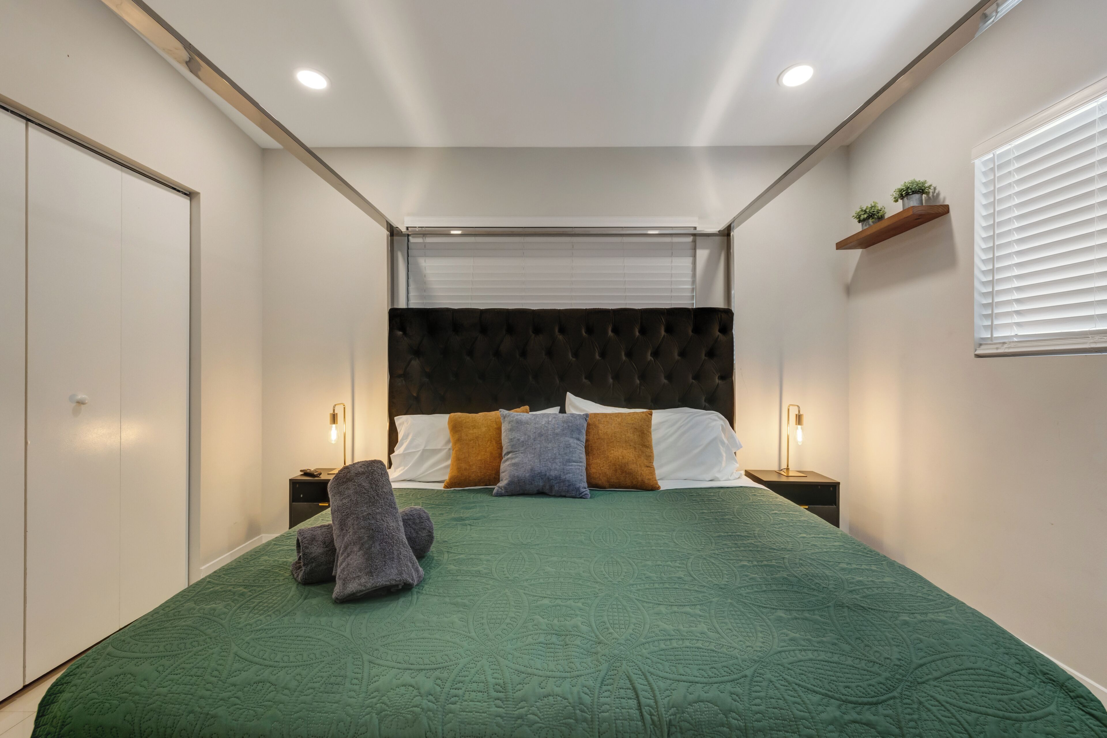 A bedroom with a green bedspread, a tufted headboard, bedside lamps, and a window with blinds.