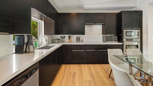 A modern kitchen with dark cabinetry, a dishwasher, a coffee maker, a microwave, and a glass dining table. - Villa Solara | Pool · Cinema · Basketball Court  (North Miami Beach)