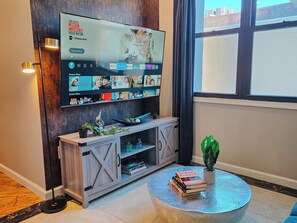 Ein Wohnzimmer mit einem an der Wand montierten Fernseher, der eine Streaming-Oberfläche anzeigt, einer Holzunterhaltungsecke mit Büchern und einem runden Couchtisch mit Büchern und einer Pflanze.