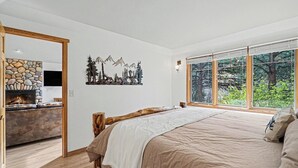 Ein Schlafzimmer mit einem großen Bett, einem Kamin aus Stein und Blick auf Bäume durch große Fenster.
