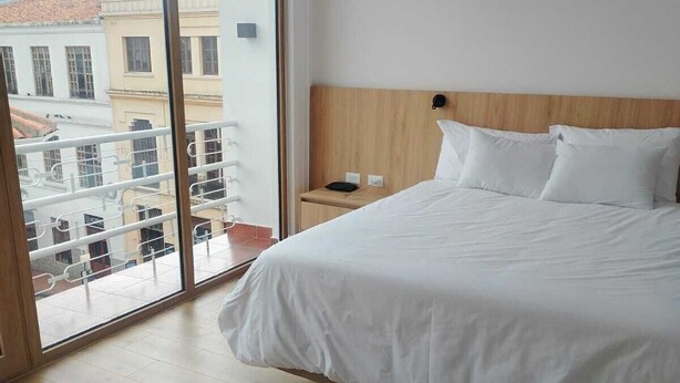 Une chambre d’hôtel avec un grand lit, une tête de lit en bois, une table de chevet et une vue sur les bâtiments, visible à travers une porte vitrée.