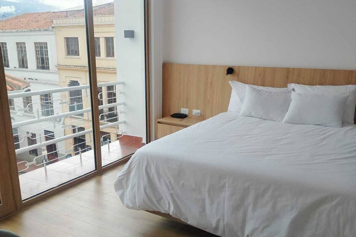 Ein Hotelzimmer mit einem großen Bett, einem hölzernen Kopfteil, einem Nachttisch und Blick auf Gebäude durch eine Glastür.