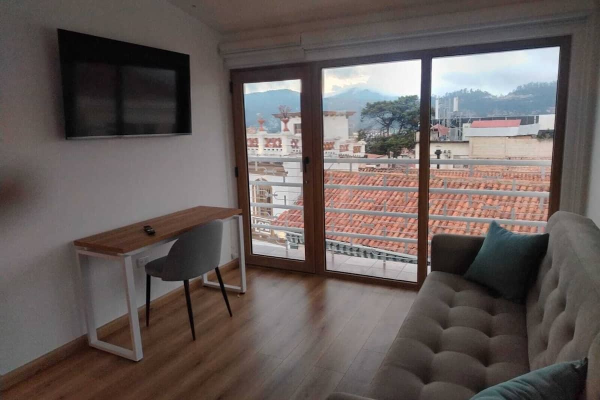 Ein Zimmer mit einem Holztisch, einem grauen Sessel, einem grauen Sofa und einer großen Schiebeglastür, die auf eine Terrasse mit roten Ziegeldächern führt.