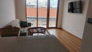 A room with a dining table, two chairs, a sofa with cushions, a TV on the wall, and a large sliding glass door leading to an outdoor area with a tiled roof. - ART DECO CUENCA (Cuenca)
