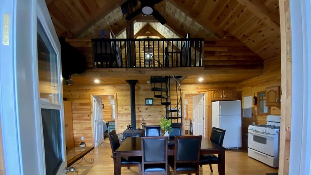 A wooden cabin interior with a dining area, kitchen, and a loft bedroom. - The Cottage on Robin Hood Lake Off Grid Adventures (Grand Bay-Westfield)