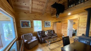 A wooden cabin interior with a fireplace, leather sofas, a dining table, and a bear mounted on the ceiling. - The Cottage on Robin Hood Lake Off Grid Adventures (Grand Bay-Westfield)