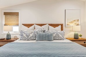 A bedroom with a large bed, multiple pillows, a wooden headboard, bedside tables with lamps, a window with a blind, and a framed picture on the wall. - Steps to Sand | Beach Home (Manhattan Beach)