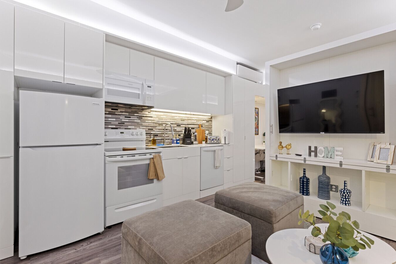 Une cuisine moderne avec des meubles blancs, un four encastré et un grand téléviseur à écran plat fixé au mur.