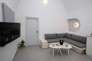 A modern living room with a flat-screen TV, a grey sofa, a small round table with drinks, and a potted plant. - Iliana s Getaway - Private Villa With Jacuzzi (Santorini)