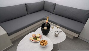 A modern living room with a grey sofa, a round coffee table, a bottle of champagne, glasses, and a plate of snacks. - Iliana s Getaway - Private Villa With Jacuzzi (Santorini)
