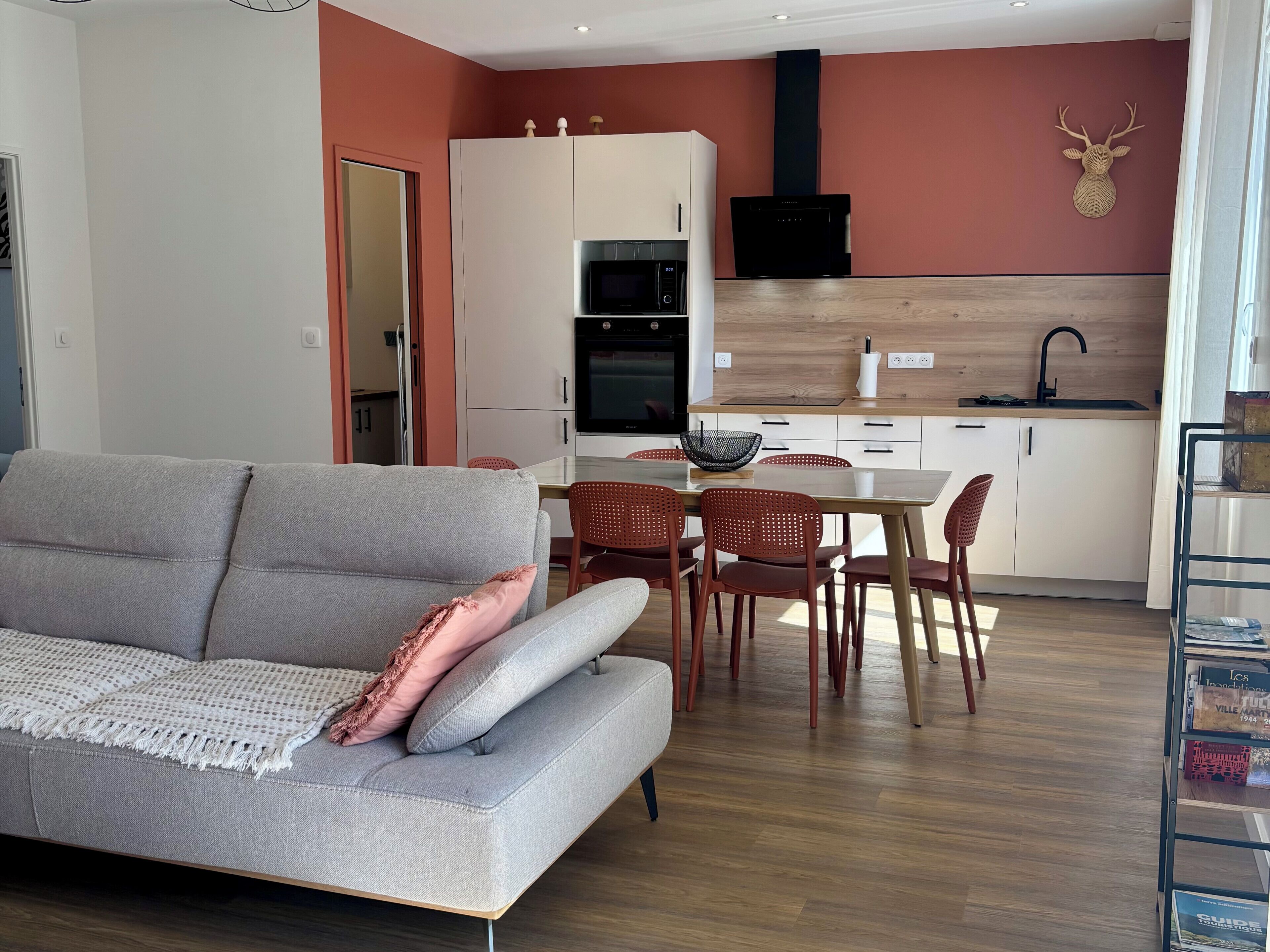 A modern living room with a grey sofa, a dining area with red chairs, a kitchen with white cabinets, and a mounted deer head on the wall.
