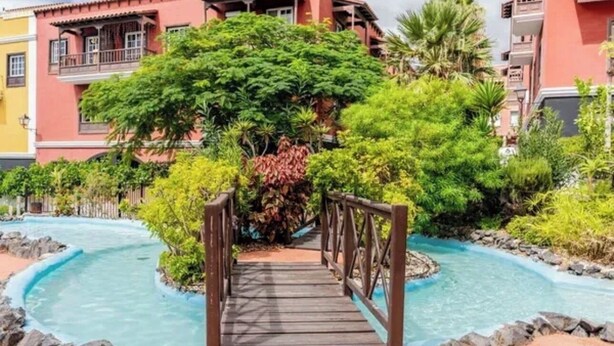 Un puente de madera que lleva a una zona de piscina rodeada de abundante vegetación y un edificio de varios pisos con balcones.