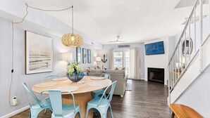 A dining area with a round table, blue chairs, a hanging light fixture, and a fireplace in the background. - Southside Seashell Escape - Ocean Block Condo with Pool (Ocean City)