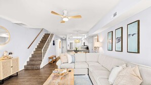 A spacious living room with a wooden coffee table, a beige sofa, a staircase, and a kitchen in the background. - Southside Seashell Escape - Ocean Block Condo with Pool (Ocean City)
