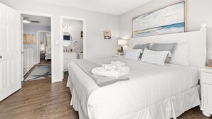 A bedroom with a large bed, a bedside table, a nightstand, a wall-mounted lamp, and a painting. - Southside Seashell Escape - Ocean Block Condo with Pool (Ocean City)
