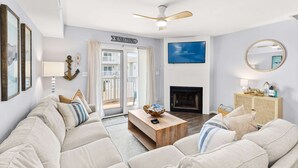 A living room with a sofa, coffee table, TV, fireplace, and a sign that reads 'SUNBATHING'. - Southside Seashell Escape - Ocean Block Condo with Pool (Ocean City)