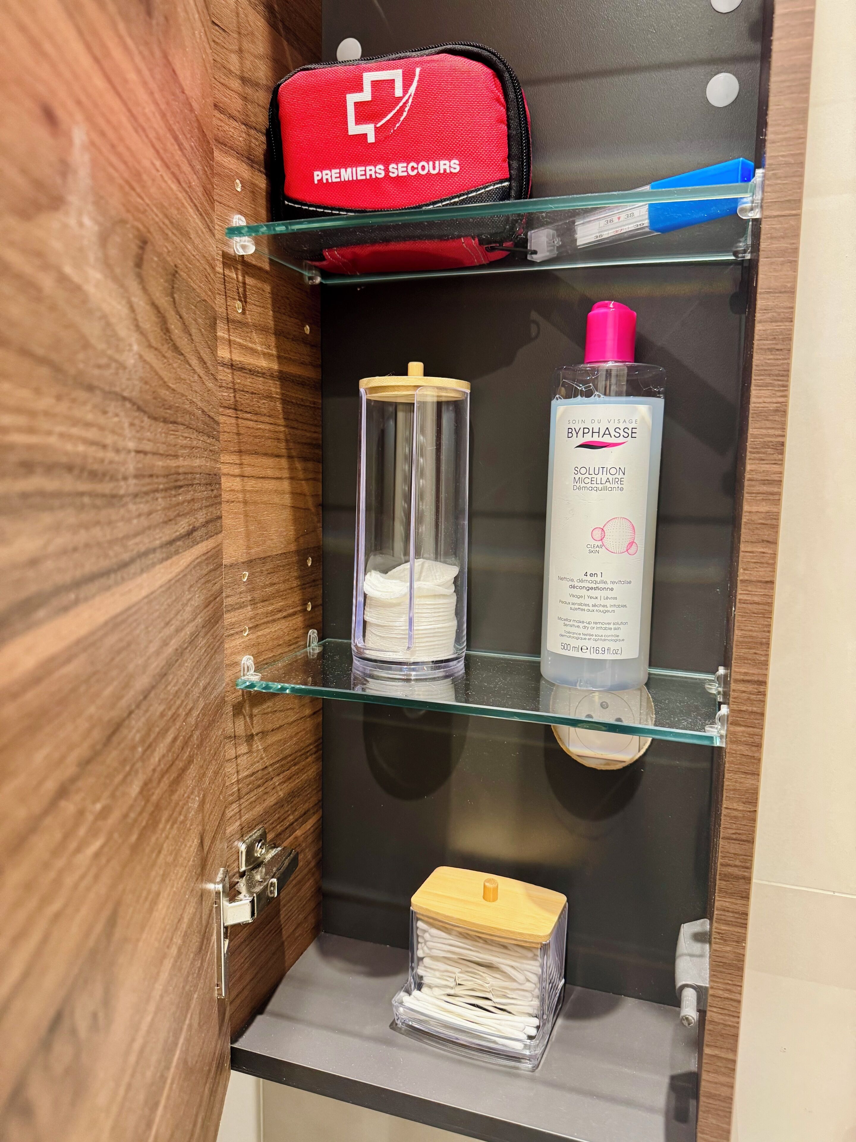 A bathroom with a glass shelf holding a red bag, a bottle of skincare product, and a container of cotton pads.