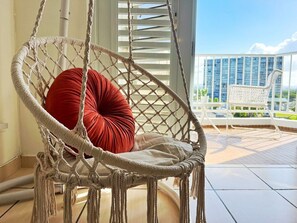 Een macramé stoel met een rode kussen, een witte stoel en een balkon met uitzicht op gebouwen.