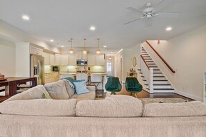 Ein geräumiges Wohnzimmer mit einer Couch, Sesseln, einem Esstisch, einer Küche im Hintergrund und einer Treppe, die zu einer weiteren Ebene führt.