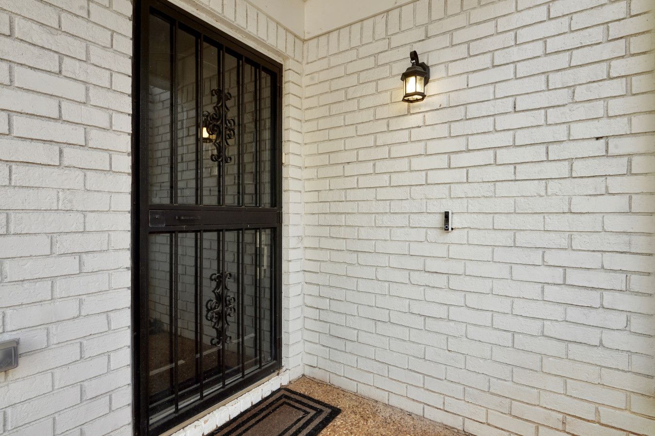 Fachada de tijolos brancos com porta de metal preta e luminária fixada na parede.