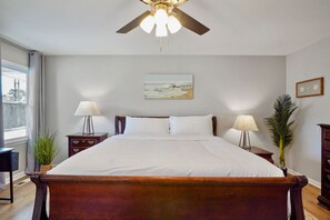 Un dormitorio con una cama grande, dos mesitas de noche con lámparas, un ventilador de techo, una ventana con cortinas, un cuadro en la pared y una planta en maceta.
