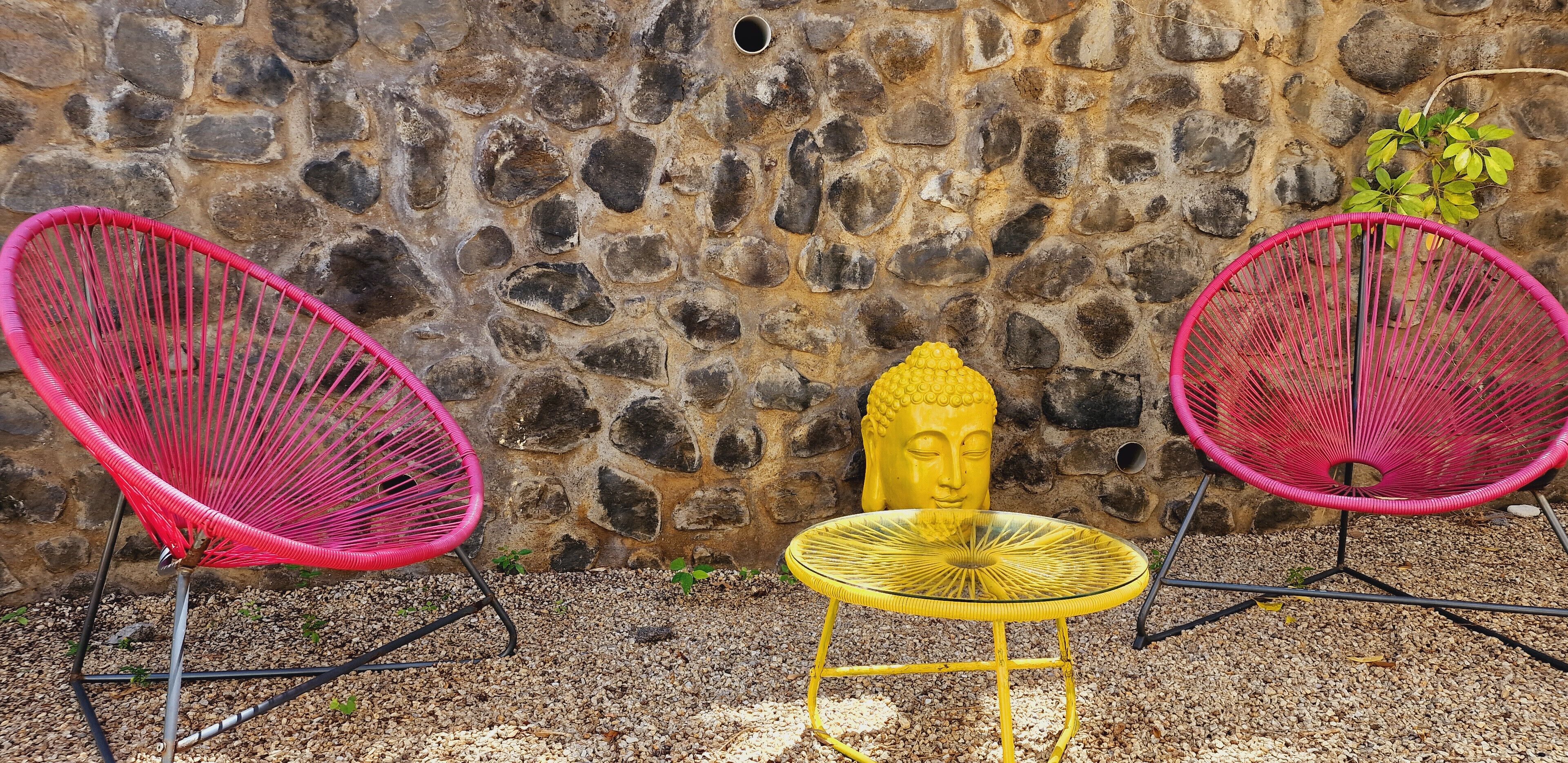Un muro in pietra, due sedie rosa, un tavolo giallo e una statua di Buddha.
