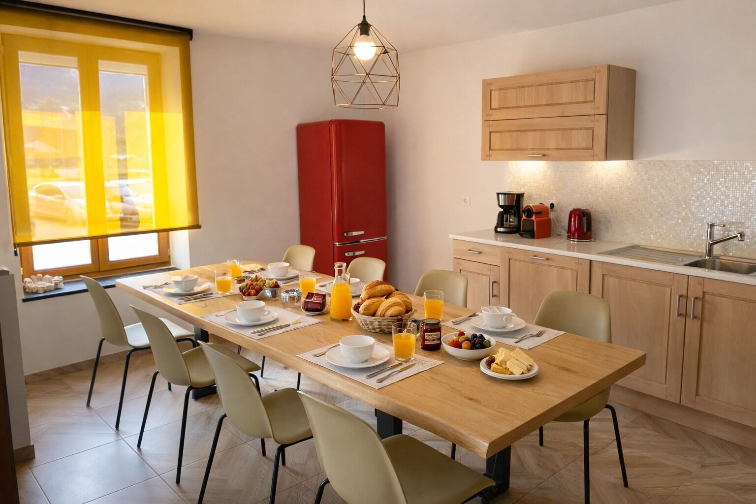 Una mesa de madera con sillas, alimentos para el desayuno, un electrodoméstico rojo y una cocina con armarios y un lavamanos.