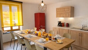 Uma mesa de madeira com cadeiras, itens de café da manhã, um aparelho vermelho e uma área de cozinha com armários e pia.