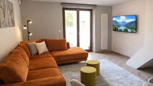Un salon comprenant un canapé d’angle, deux poufs ronds, un téléviseur à écran plat et un radiateur.