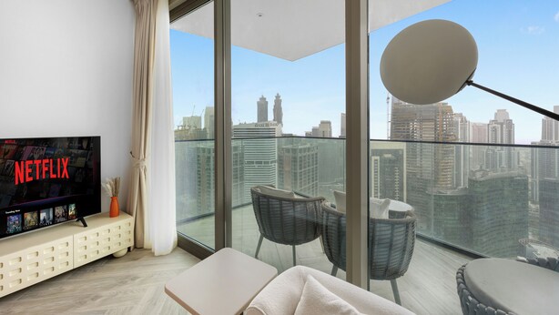 A modern living room with a large TV displaying 'NETFLIX,' a sofa, two chairs, a small table, and a balcony with a city view. - Luxury 1BR l Burj Khalifa View in Business Bay (Dubai)