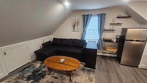 A compact living space with a black sectional sofa, a wooden coffee table, a built-in kitchen with a refrigerator, and a window with blue curtains.