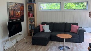 Ein modernes Wohnzimmer mit einem Ecksofa, einem Flachbildfernseher auf einem Schrank, einem Bücherregal und zwei Fenstern mit Blick auf Grünflächen.