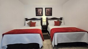 A hotel room with two beds, each with a red blanket, white bedding, and a headboard. There is a nightstand between the beds with a lamp, and two framed pictures on the wall above.