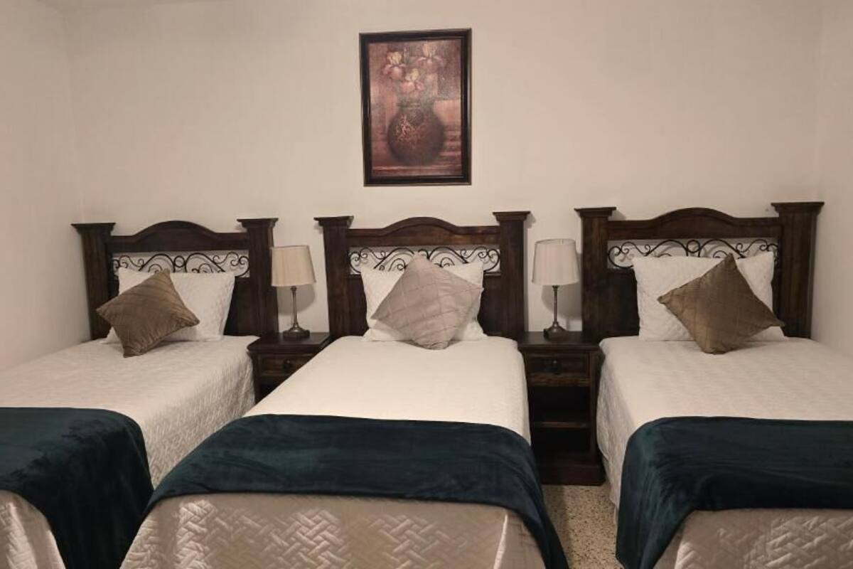 Una habitación con tres camas individuales, cada una con cabecero de madera, una mesita de noche y una lámpara.