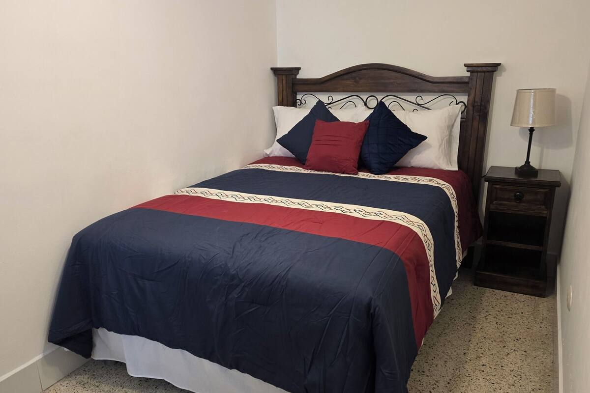 Una cama con colcha azul oscuro y roja, un cabecero de madera y una mesita de noche con lámpara.