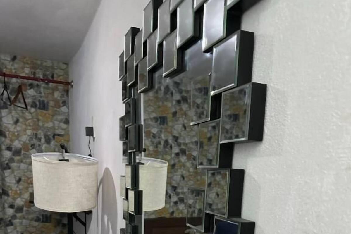 A room with a wall-mounted mirror, a bedside lamp, a bed with patterned bedding, and a wooden floor.