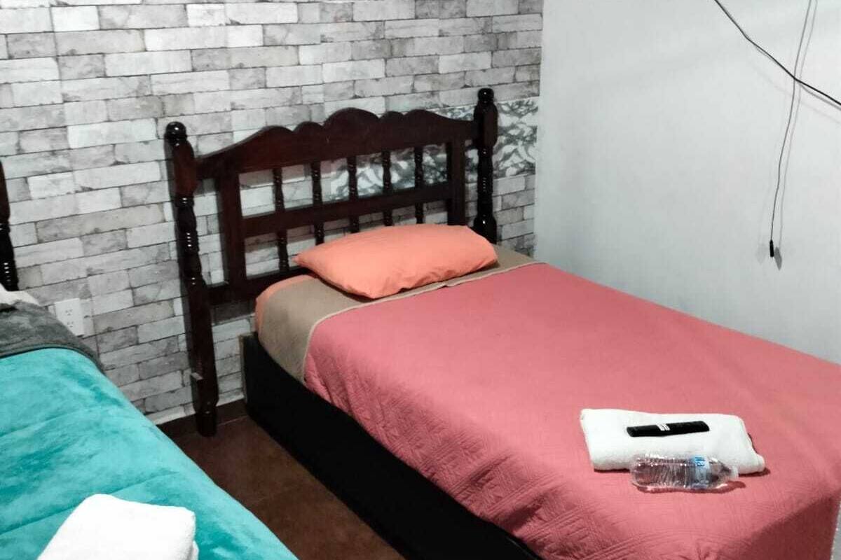 A room with two beds, one with a teal bedspread and the other with a red bedspread, a wooden headboard, a brick-patterned wall, and a mounted TV.