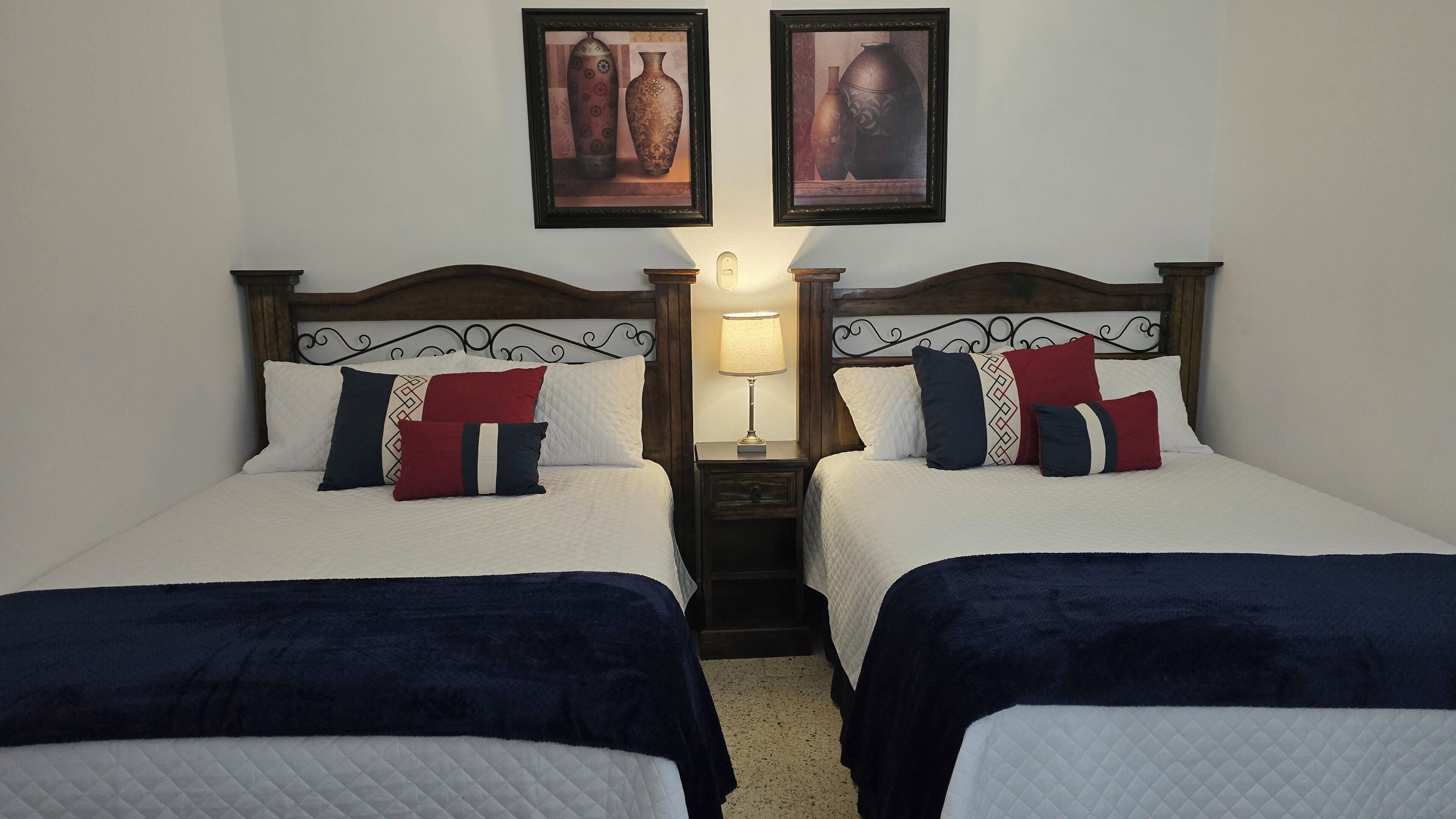 A hotel room with two beds, a nightstand with a lamp, and two framed pictures on the wall.