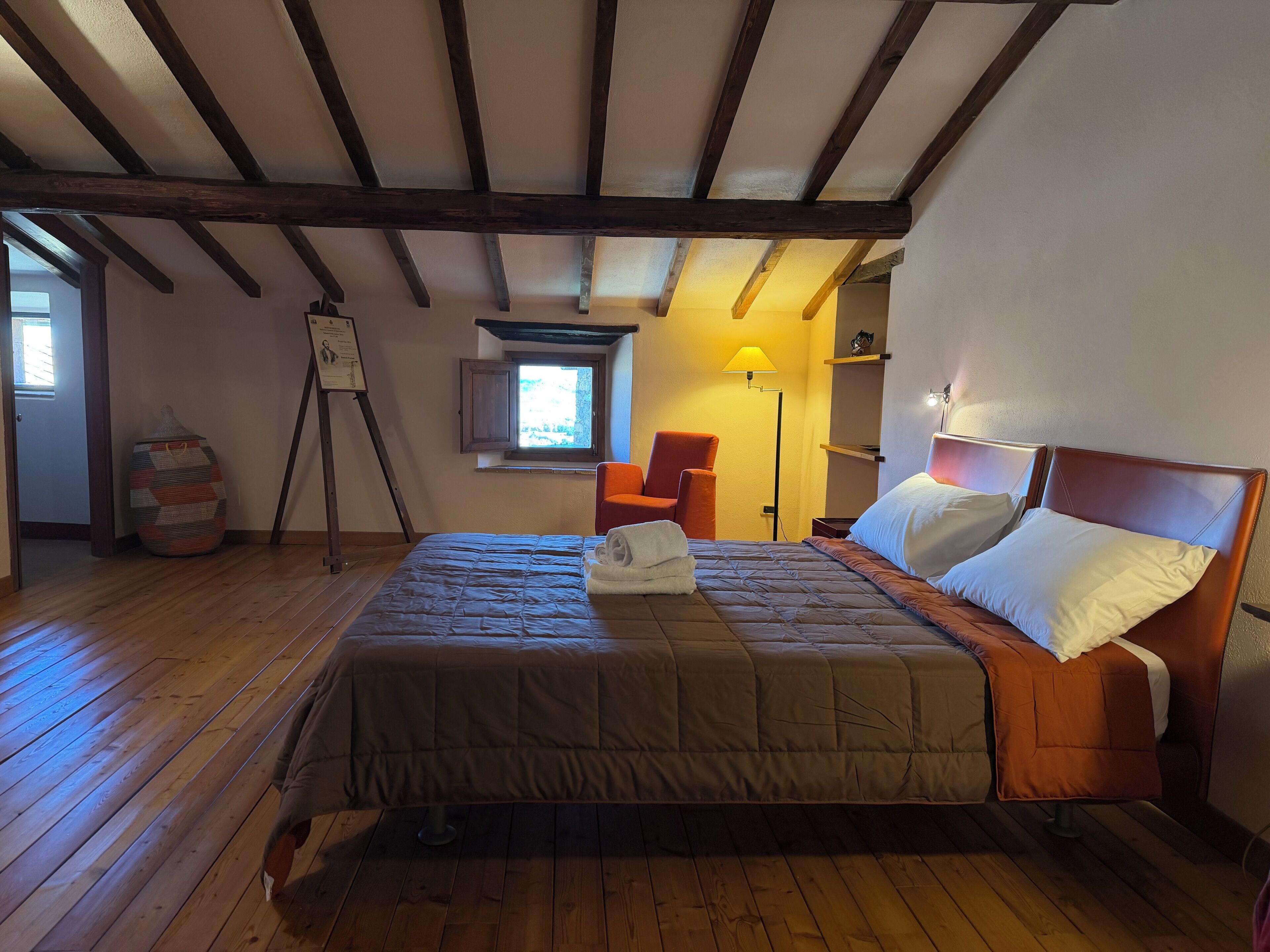 Une chambre avec un plancher en bois, un lit avec une tête de lit, une chaise, une lampe et une fenêtre.