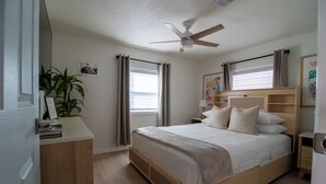 Een slaapkamer met een bed, een plafondventilator, een nachtkastje, een plant en een raam met gordijnen.