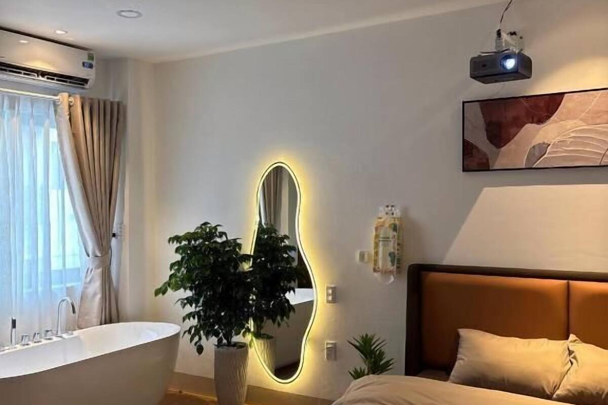 Una habitación de hotel moderna con bañera, un espejo grande con iluminación LED, una cama con ropa de cama color beige y un proyector en el techo.