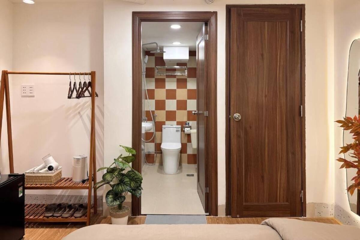 Una habitación con una cama, una puerta de madera, un baño con pared a cuadros y una planta.