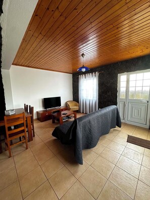 A living room with a wooden ceiling, a television, a sofa, a dining table, and a stone wall.